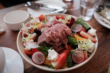 Top view of a colorful fresh salad bowl with strawberries, citrus, radish, leafy greens, cheese and shredded meat, served on a wooden table with plates and cutlery. Healthy vibrant meal.