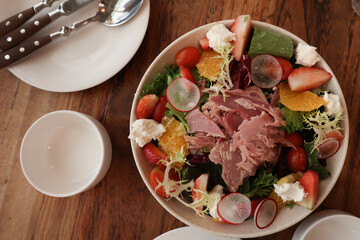 Top view of a colorful fresh salad bowl with strawberries, citrus, radish, leafy greens, cheese and shredded meat, served on a wooden table with plates and cutlery. Healthy vibrant meal.