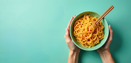 Hands hold a bowl of noodles with chopsticks. Yellow pasta is ready for eating. Food photography on aqua menthe background shows simple meal in bowl from above.