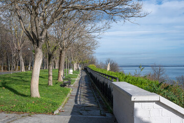 A quiet park path bordered by leafless trees and a white wall overlooks a calm body of water in Burgas, Bulgaria