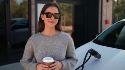 A stylish woman in sunglasses holds a coffee cup near a charging electric car Stock video
