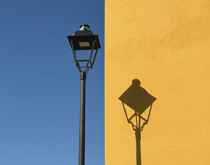 alte Stra&szlig;enlaterne vor blauem Himmel wirft ihren Schatten auf eine gelbe Hauswand