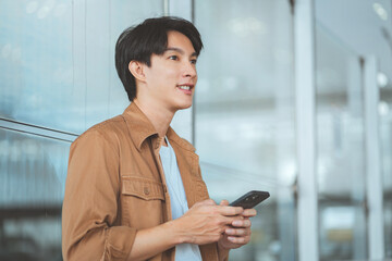 Attractive young asian man wearing casual clothes using smartphone near airport entrance &ndash; browsing internet, checking messages and staying connected while traveling with digital lifestyle