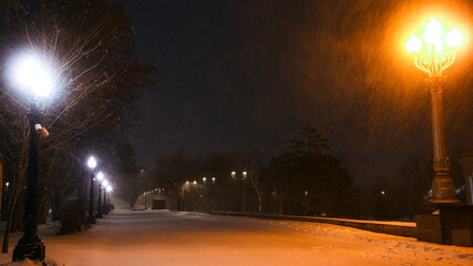 Fabulous view of the snow-covered city park at night, with trees in the snow, cozy warm light of lanterns.