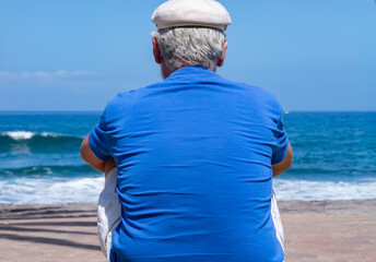Senior man sitting alone facing the ocean enjoying a peaceful moment by the seaside sitting near the beach with waves and clear blue sky in the background. Relaxation and retirement