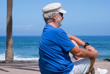 Senior man sitting alone facing the ocean enjoying a peaceful moment by the seaside sitting near the beach with waves and clear blue sky in the background. Relaxation and retirement