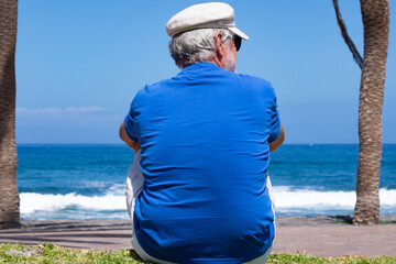 Senior man sitting alone facing the ocean enjoying a peaceful moment by the seaside sitting on the grass near palm trees with waves and clear blue sky in the background. Relaxation and retirement