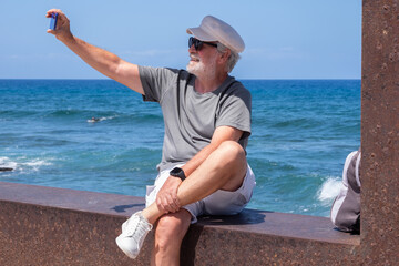 Senior man smiling taking a selfie by smartphone enjoying a peaceful moment by the seaside sitting near the beach with waves and clear blue sky in the background. Relaxation and retirement