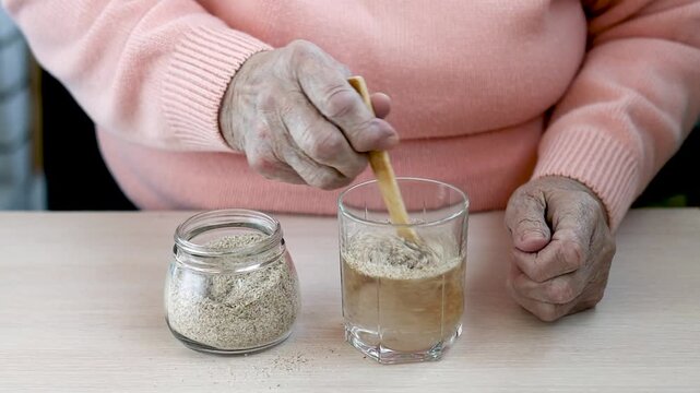 the hands of an old woman pour psyllium husks into a glass of water and mix