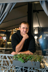 A thoughtful man in a black hoodie leans on a decorative railing with a planter at night