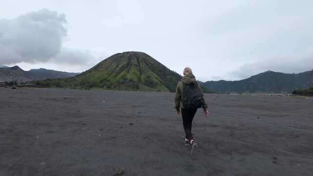 Woman walks alone across black volcanic sand toward lush Mount Batok and smoking Mount Bromo in Indonesia. Scenic travel moment filled with solitude, nature, and adventure.
