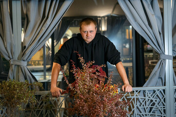 A man in a black hoodie stares intensely while leaning on a decorative railing with colorful foliage,
