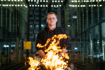 A man in a black hoodie stands centered, illuminated by the intense flames of a bonfire in the foreground