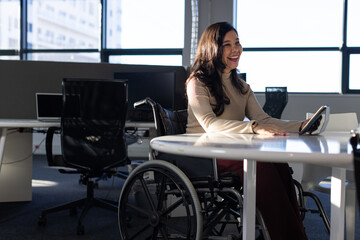 Woman in wheelchair smiling while holding tablet at white table in office workspace, copy space