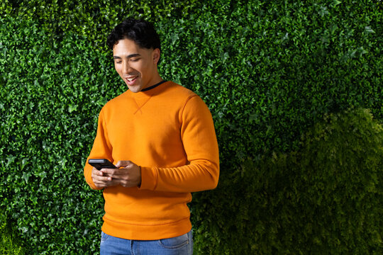 Asian man standing by hedge in garden smiling while holding smartphone wearing orange sweatshirt