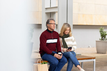 Senior couple sitting on bench outside building, holding coffee beside bag and using smartphone