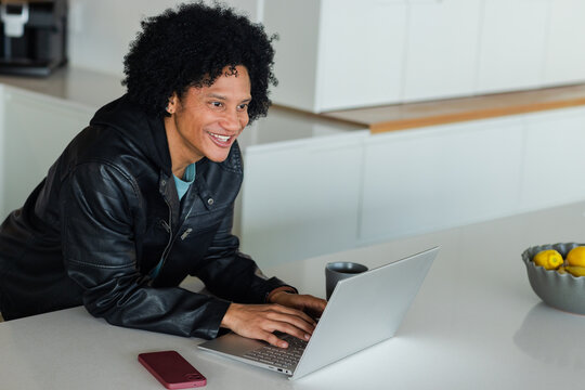 African American man leaning over kitchen island, smiling and typing on silver laptop near gray mug