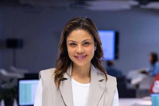 Mid adult Hispanic woman smiling while standing in office with computer monitors and plant