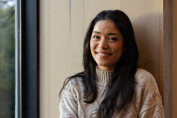 Woman sitting by window in room against wooden paneled wall, wearing knit sweater and hoop earrings