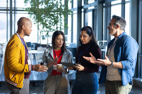 Diverse colleagues in business attire discussing in office lounge with coffee cups, tablet and IDs