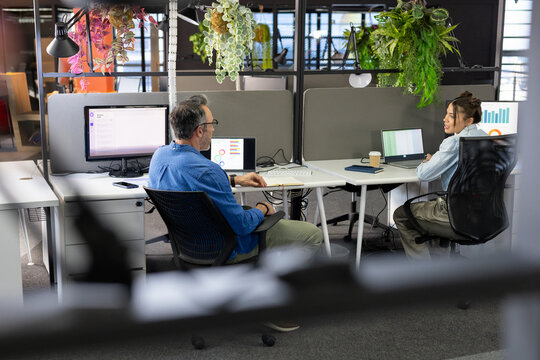 Diverse coworkers consulting over spreadsheet on laptop, chart on monitor at open-plan office desks