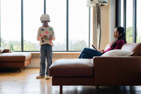Indian mother and daughter using VR headset and tablet on leather sofas in living room