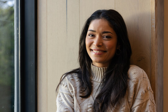 Woman sitting by window in room against wooden paneled wall, wearing knit sweater and hoop earrings