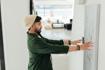 Indian man attaching canvas painting to wall with bracket in living area of home, copy space