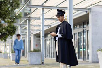 Male graduate wearing mortarboard and gown checking smartphone with smartwatch on campus walkway