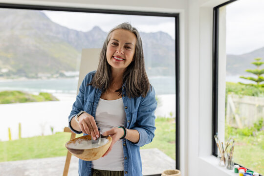 Woman stirring paint mixture in wooden bowl in coastal art studio with paintbrushes on windowsill
