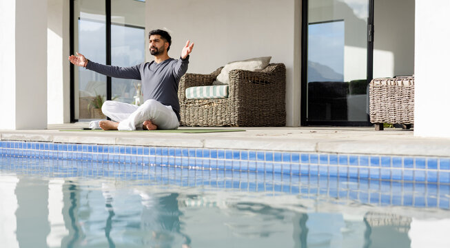 Indian adult male practicing yoga on green mat at poolside patio with wicker sofa cushions