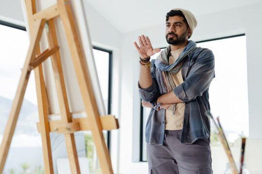 Indian man painting canvas at art studio by black-framed windows, holding paintbrushes beside easel