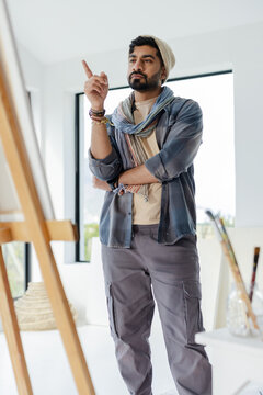 Indian male artist standing in art studio pointing at blank canvas by easel with paintbrush jars