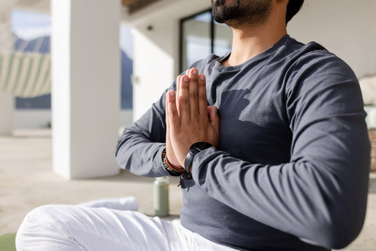 Indian man kneeling on green yoga mat on terrace, practicing yoga with smartwatch and bottle