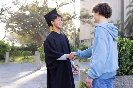 Diverse male friends shaking hands on campus path with capped graduate holding diploma scroll