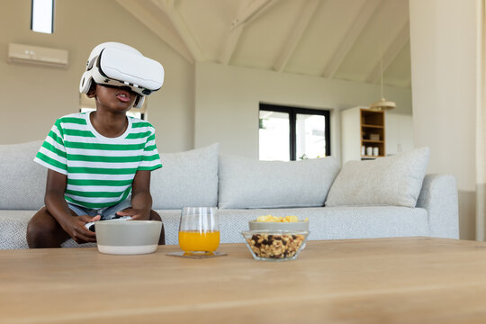 Child wearing VR headset holding remote sitting on sofa beside coffee table in modern living room