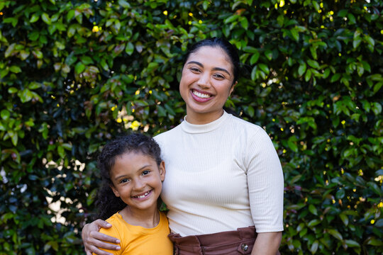 Mother and daughter standing in garden wearing cream top brown bottoms yellow shirt against hedge