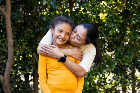 Diverse mother and daughter hugging and kissing while standing in garden with black smartwatch - Powered by Adobe