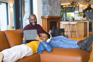 African American father and son laughing while using smartphone and tablet on sofa at home