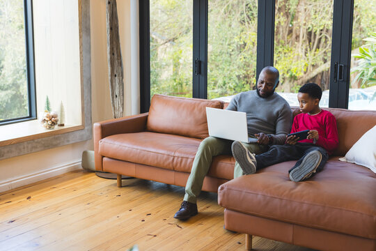 African American father and son using laptop and tablet on sectional sofa in living room