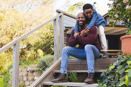 African American father and son sitting on patio steps near cable railing hugging near planter