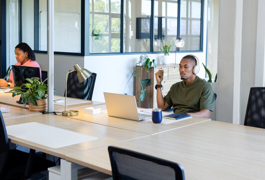 Coworkers sitting at open office desks typing on laptop with headphones and coffee mug, copy space