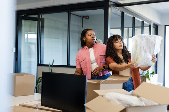 African American female coworkers unpacking bubble-wrapped decor from boxes in office with monitor
