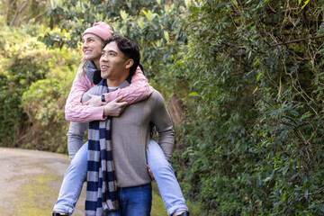 Diverse couple giving piggyback ride along paved park trail wearing striped scarf and pink beanie