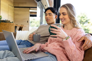 Diverse couple holding mugs while using silver laptop and sitting on sofa in modern living room