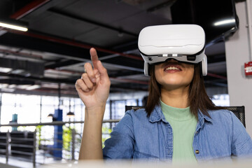 Woman sitting wearing white VR headset and interacting with virtual controls in open-plan office