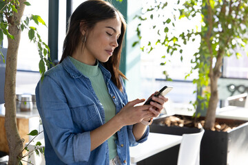 Asian Indian woman holding smartphone and scrolling messages in office by ficus trees, copy space