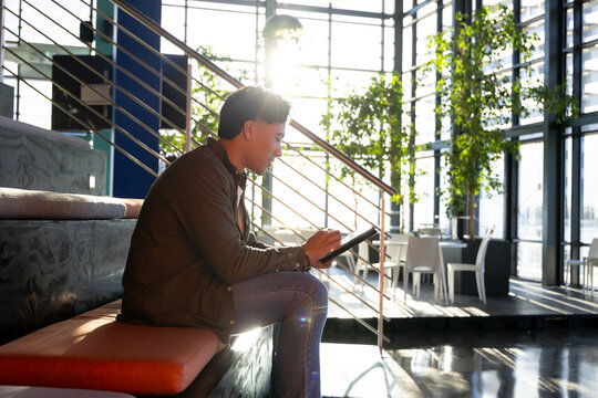 Man sketching on tablet with stylus while sitting on cushioned bench by metal staircase and windows - Powered by Adobe