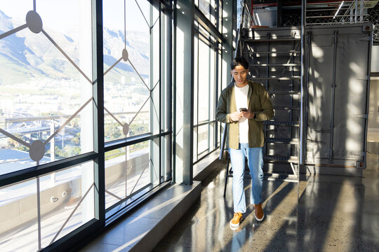 Asian man walking along corridor using smartphone past windows and container staircase, copy space