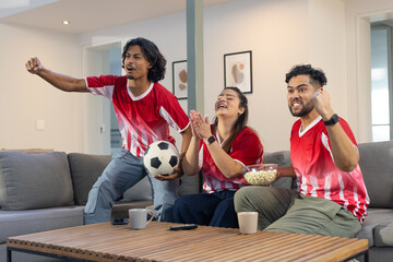 Cheering friends wearing red jerseys sitting on couch in living room with soccer ball and popcorn